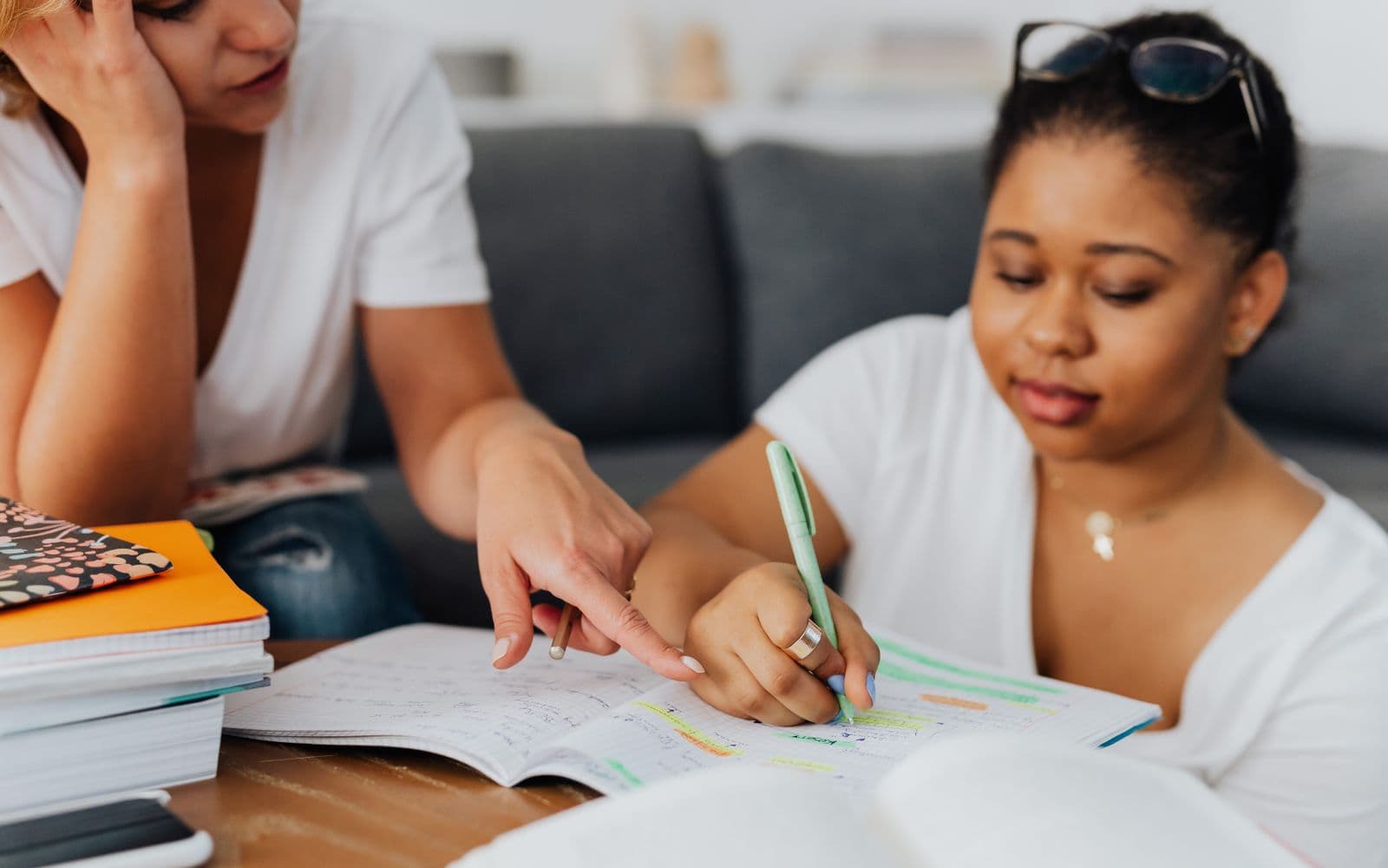 Two women sit together on a couch looking at an open notebook, one pointing at the page while the other writes with a pen.